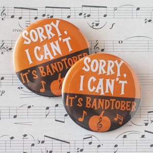 May include: Two round orange and black buttons with the text "SORRY, I CAN'T It's BANDTOBER" in white. The buttons feature a pumpkin and musical notes. The background is sheet music.