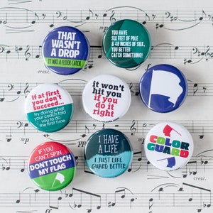 May include: Ten colorful round buttons with various sayings related to color guard. The buttons feature green, blue, red, white, and black colors. The text on the buttons includes: "That wasn't a drop", "It was a floor catch", "If at first you don't succeed...", "Try doing what your coach told you to do the first time", "You have six feet of pole & 48 inches of silk, you better catch something!", "It won't hit you if you do it right", "If you can't spin don't touch my flag", "I have a life I just like guard better", "Color Guard".