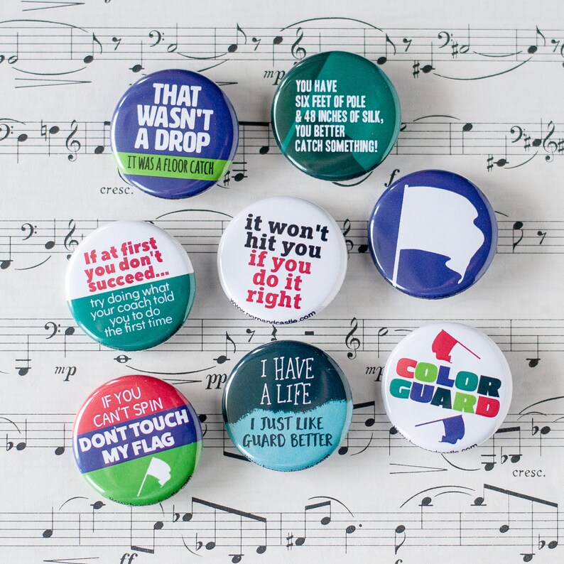 May include: Ten colorful round buttons with various sayings related to color guard. The buttons feature green, blue, red, white, and black colors. The text on the buttons includes: "That wasn't a drop", "It was a floor catch", "If at first you don't succeed...", "Try doing what your coach told you to do the first time", "You have six feet of pole & 48 inches of silk, you better catch something!", "It won't hit you if you do it right", "If you can't spin don't touch my flag", "I have a life I just like guard better", "Color Guard".