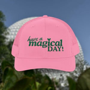 May include: A pink trucker cap with a mesh back and a curved peak. The front of the cap has the embroidered phrase "have a magical DAY!" in green script. The cap is set against a blurred background.