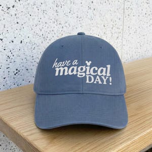 May include: A dusty blue baseball cap featuring the white embroidered text "have a magical DAY!" with a small mouse ear icon above the word magical. The cap has a structured crown, a curved brim, and a classic six-panel design with top stitching.