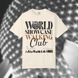 May include: A cream-coloured short-sleeve t-shirt featuring black and tan text that reads WORLD SHOWCASE WALKING CLUB, SEE THE WORLD IN 1.2 MILES, and a graphic of various building silhouettes. The letter O in WORLD is replaced by a textured sphere.