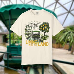 May include: Cream-colored t-shirt with a graphic of a boat ride through a greenhouse and an orange tree. The text "ONLY THEN, WILL WE TRULY BE" is arched above the image, and "LIVING WITH THE LAND" is below. The shirt is a crew neck.
