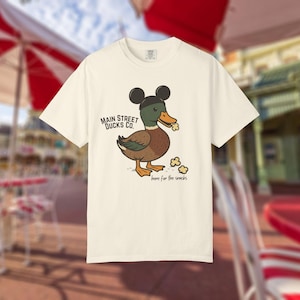 Może przedstawiać: Kremowa koszulka z kreskówkową kaczką w czapce Myszki Miki i popcornem. Koszulka ma napis "Main Street Ducks Co." i "here for the snacks". Kaczka jest brązowa, zielona i pomarańczowa.