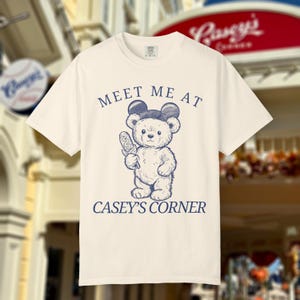 May include: Cream-colored t-shirt featuring a navy blue illustration of a teddy bear wearing mouse ears and holding a corn dog. The text "MEET ME AT CASEY'S CORNER" is printed above and below the bear.