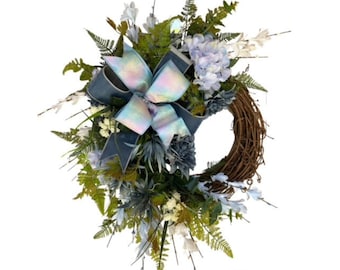 May include: A decorative wreath featuring a natural brown grapevine base, adorned with a large iridescent bow and an assortment of artificial flowers and greenery. The wreath includes blue, white, and green elements.