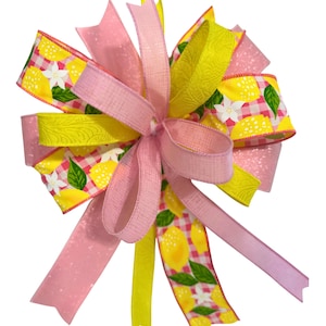 May include: A decorative bow featuring a lemon theme. The bow has multiple layers of pink and yellow ribbons. Some ribbons have a lemon and floral pattern, while others are solid colors or have a glittery finish. The bow is suitable for various craft projects.