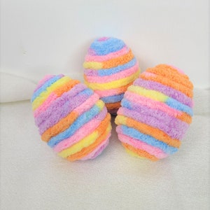May include: Three colorful Easter eggs made of yarn. The eggs are striped with pink, blue, yellow, and orange yarn.