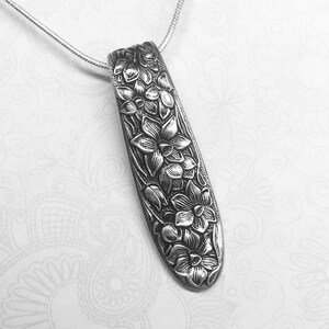 May include: A silver necklace with a pendant shaped like a long, narrow leaf with a floral design. The pendant is made from a repurposed vintage spoon.