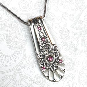 May include: A silver-toned pendant necklace featuring a repurposed spoon handle with an embossed floral pattern. The metal surface is decorated with several small, round, bright pink crystals embedded within the center of the sculpted flowers.