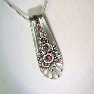 May include: A silver spoon pendant necklace with floral detailing and pink gemstone accents. The spoon bowl is adorned with embossed flowers and leaves, and the pendant hangs from a delicate silver chain. The pendant measures approximately 3 inches in length.