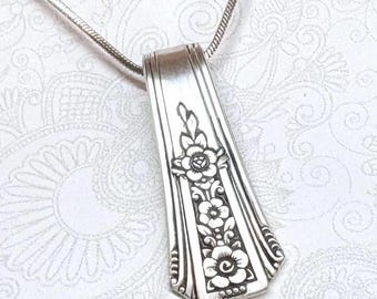 May include: A silver-toned pendant necklace featuring a flat, tapered metal handle with an embossed floral pattern of three stacked flowers and leaves. The pendant is suspended from a thin, flexible silver-toned snake chain necklace.