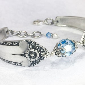 May include: A silver spoon bracelet with a light blue crystal bead and a silver chain. The spoon has a floral design.