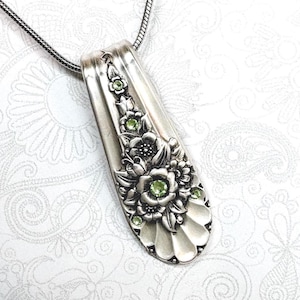 May include: A silver-toned pendant crafted from a repurposed spoon handle, featuring an embossed floral pattern and inlaid with small, round, light green crystals. The pendant is suspended from a thin, flexible silver-toned snake chain necklace.