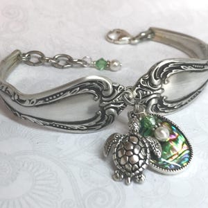 May include: A silver-toned bracelet crafted from a repurposed ornate spoon handle featuring intricate embossed detailing. The bracelet includes a dangling charm of a sea turtle, a small white pearl bead, and an oval iridescent abalone shell.