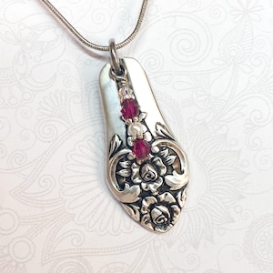May include: A silver-toned pendant necklace featuring a teardrop-shaped metal piece with embossed floral patterns. The pendant is accented with a vertical row of two deep pink faceted beads and one central white pearl bead, suspended on a thin chain.