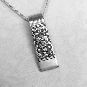May include: A silver pendant necklace with a rectangular pendant. The pendant has an ornate floral design with a detailed pattern. The necklace chain is a thin, delicate silver chain. The background is a white, patterned surface.