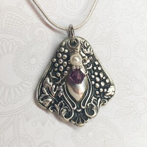 May include: A silver-toned pendant necklace featuring an ornate, embossed metal design of grape clusters and leaves. The pendant is accented with a single white pearl bead and a faceted purple crystal bead, suspended from a thin silver-toned chain.