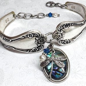 May include: A silver-toned bracelet crafted from a bent spoon handle with floral engravings, featuring a lobster clasp and an extender chain. A pendant hangs from the centre, showcasing an oval abalone shell inlay and a metal dragonfly charm.