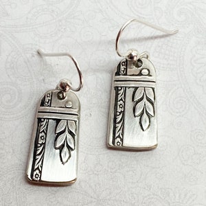 May include: A pair of silver-toned drop earrings featuring a rectangular shape with rounded top edges. Each earring displays an engraved vertical floral border and a central leaf motif, suspended from a simple silver-toned hook wire finding.