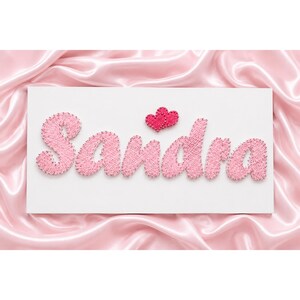 May include: A white rectangular sign with the name "Sandra" spelled out in pink string art. Two small pink hearts are above the name. The background is a pink satin fabric.