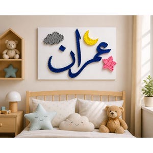 Könnte beinhalten: Ein Kinderzimmer mit einem Holzbett und einem Nachttisch. Ein Wandbehang zeigt den Namen "Imran" in blauer Fadengrafik, mit einem gelben Halbmond und einem rosa Stern. Weiche Kissen und Teddybären schaffen eine warme und einladende Atmosphäre.