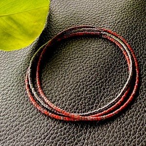 May include: Three beaded bracelets in shades of red and black are coiled on a textured black surface. The bracelets are made of small, evenly spaced beads. Green leaves and a stem are in the upper left corner.