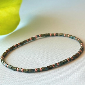 May include: An olive green and copper beaded bracelet. The bracelet features small, square beads in shades of green and copper-colored round beads. The bracelet is displayed on a light blue surface.