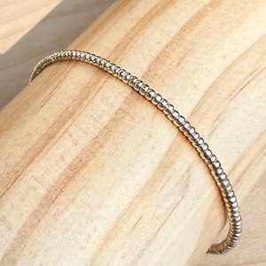 May include: A silver beaded bracelet with a flexible design. The bracelet is made of small, round beads strung together, creating a textured surface. The bracelet is shown on a wooden surface.