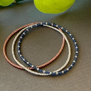 May include: Three beaded bracelets in copper, silver, and navy blue. The bracelets are made of small, round beads strung together to form a flexible band. The bracelets are stacked on top of each other.