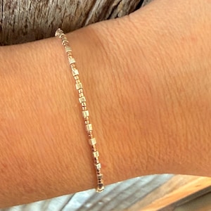 May include: A delicate bracelet composed of small, rectangular and round beads in gold and copper tones. The bracelet is worn on a wrist, showcasing its minimalist design and subtle elegance. The beads are strung together, creating a refined accessory.