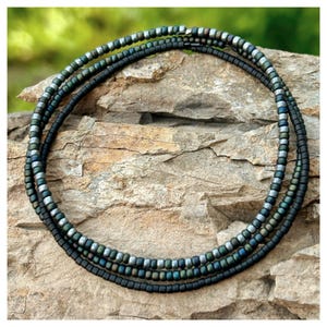 May include: Three layered beaded necklace with a mix of black, silver, and teal beads. The necklace is displayed on a textured stone surface, with a blurred green background.