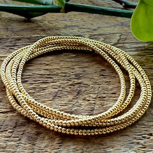May include: A stack of gold beaded bracelets. The bracelets are made of small, round, gold-colored beads strung together. The bracelets are arranged in a circular shape on a wooden surface.