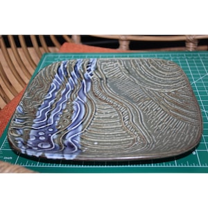 May include: Square ceramic plate with a textured surface. The plate features a design with blue and white wave-like patterns on one side and a textured, earthy-toned pattern on the other. The plate is approximately 8 inches square.