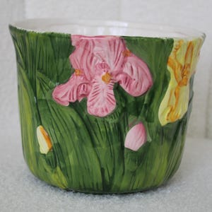 May include: A ceramic planter with a white interior and a green exterior featuring embossed iris flowers. The planter has pink and yellow iris flowers with green leaves. The planter is a decorative item for indoor or outdoor use.