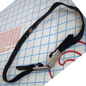 May include: A black belt with a rectangular silver buckle encrusted with small, sparkling rhinestones. The belt is made of a smooth, dark fabric and features a small label with the letters "MM".