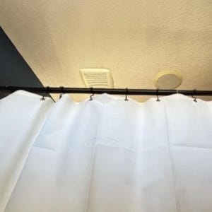 May include: A white shower curtain suspended from a black rod with black hooks. The curtain has gentle folds. Above the rod, a white vent and a circular light fitting are visible against a beige ceiling.