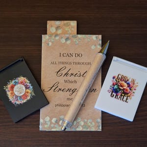May include: An assortment of inspirational items, including a pen and two small notebooks. The pen has a metallic tip and a beige barrel. One notebook is black with a floral design, and the other is white with a similar floral design. The central card reads "I can do all things through Christ".