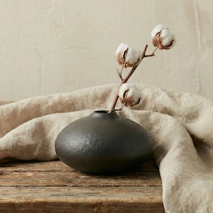 May include: A small, dark grey ceramic vase with a textured surface holds a sprig of cotton blossoms. The vase sits on a wooden surface, with a beige linen cloth draped around it. The cotton blossoms have white petals and brown stems.