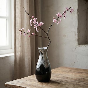 Puede incluir: Un jarrón de cerámica en blanco y negro con un borde ondulado sostiene una rama floreciente con pequeñas flores rosas. El jarrón está sobre una mesa de madera rústica cerca de una ventana con una cortina beige.