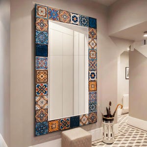 Puede incluir: Espejo rectangular grande con un borde decorativo de azulejos estampados coloridos en tonos azules, naranjas y blancos. El espejo está montado en una pared gris en un pasillo. Un pequeño otomán cuadrado beige se encuentra debajo.