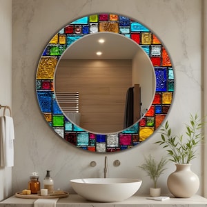 May include: A round wall mirror featuring a decorative frame composed of multi-coloured, textured glass tiles in shades of blue, red, yellow, green, and clear. The reflective surface shows a bathroom interior with a sink and vanity accessories.