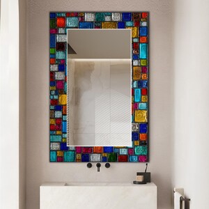 May include: A rectangular wall mirror featuring a decorative frame composed of small, multi-coloured glass tiles in shades of blue, red, yellow, green, and silver. The glass tiles have a textured, translucent appearance arranged in a mosaic pattern.
