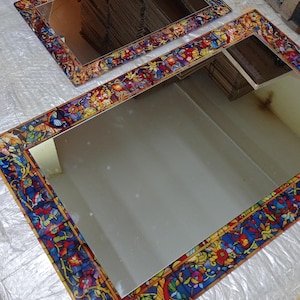 K&ouml;nnte beinhalten: Zwei rechteckige Spiegel mit farbenfrohen R&auml;ndern im Glasmalerei-Stil. Die R&auml;nder weisen eine lebendige Mischung aus roten, blauen, gelben und gr&uuml;nen floralen und abstrakten Designs auf. Die Spiegel liegen auf einer reflektierenden Oberfl&auml;che.