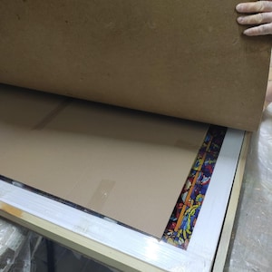 K&ouml;nnte beinhalten: Ein rechteckiges Buntglasfenster mit einem farbenfrohen Blumendesign ist teilweise in einem Holzrahmen sichtbar. Das Panel wird durch Pappe und eine Holzkiste gesch&uuml;tzt. Der Rahmen ist hell.