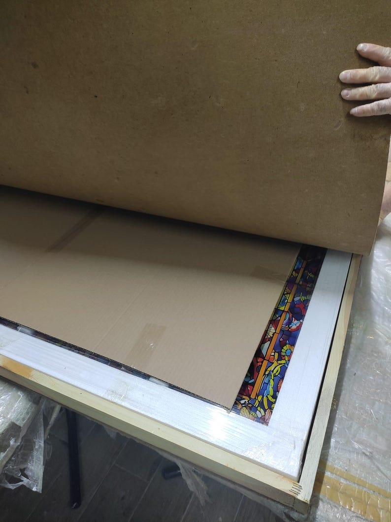 K&ouml;nnte beinhalten: Ein rechteckiges Buntglasfenster mit einem farbenfrohen Blumendesign ist teilweise in einem Holzrahmen sichtbar. Das Panel wird durch Pappe und eine Holzkiste gesch&uuml;tzt. Der Rahmen ist hell.