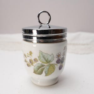 May include: A white ceramic egg cup with a silver-colored lid and handle. The cup is decorated with a floral pattern of green leaves and purple berries. A classic kitchenware item.