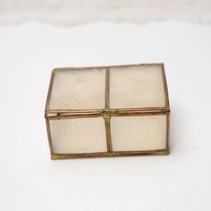 Puede incluir: Pequeña caja rectangular con tapa abisagrada. La caja presenta una parte superior transparente, similar al vidrio, dividida en cuatro secciones, enmarcada por un metal fino de color dorado. La caja se utiliza probablemente para guardar objetos pequeños, como joyas o recuerdos.