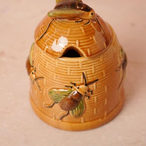 May include: A ceramic honey pot shaped like a traditional beehive, featuring a golden-brown woven texture. The lid is topped with a sculpted bee, and additional bees are molded onto the sides. It includes a small notch for a honey dipper.