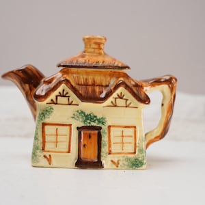 Puede incluir: Una tetera de cerámica con forma de cabaña. La tetera presenta un tejado de paja, una puerta y dos ventanas. La paleta de colores incluye acentos beige, marrón y verde. El asa y el pico también son marrones.